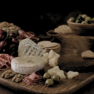 A moody close-up of a wooden board with cheeses, crackers, salami, grapes, olives, and walnuts — a fine spread, before Jam Vino